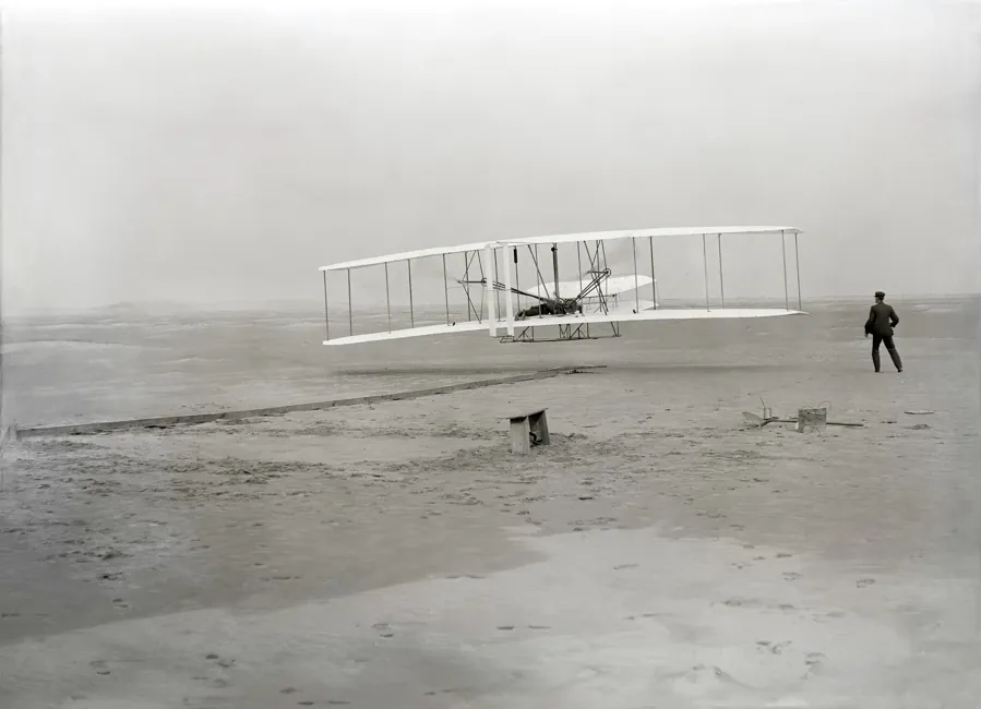 O primeiro voo dos Wright, em 17 de dezembro de 1903. Orville pilota, Wilbur corre ao lado. Foto: John T. Daniels, domínio público